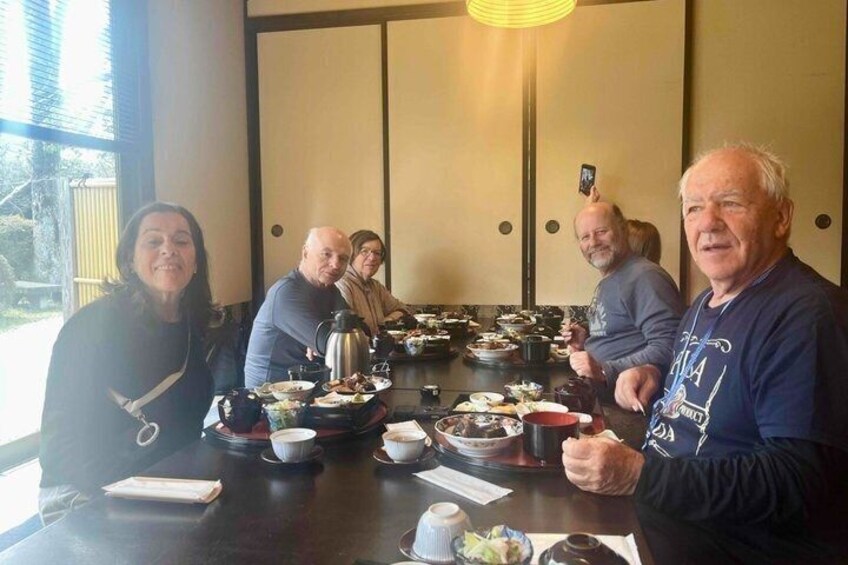 A group of people seated around a table with various dishes, a kettle, and a smartphone in use. 3