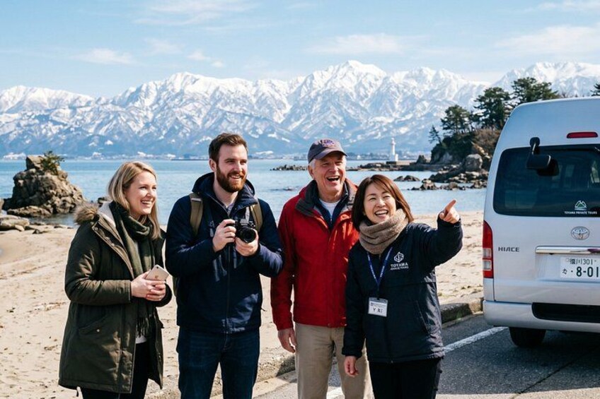 A group of people standing on a beach with a vehicle and snow-capped mountains in the background. 1