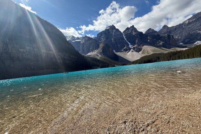 A clear mountain lake with rocky shore, surrounded by forested mountains under a partly cloudy sky. 2