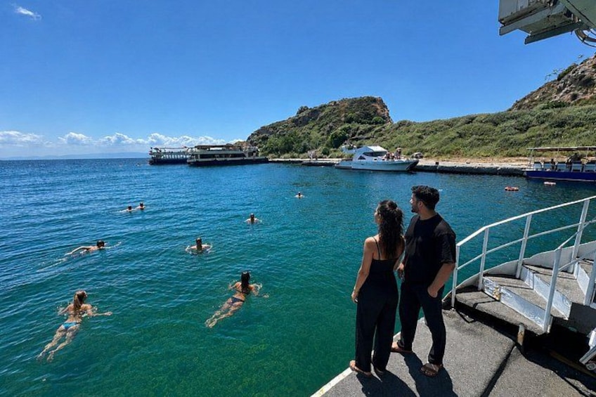 A couple standing on a dock overlooking a clear blue sea with swimmers in the water and boats in the background. 1