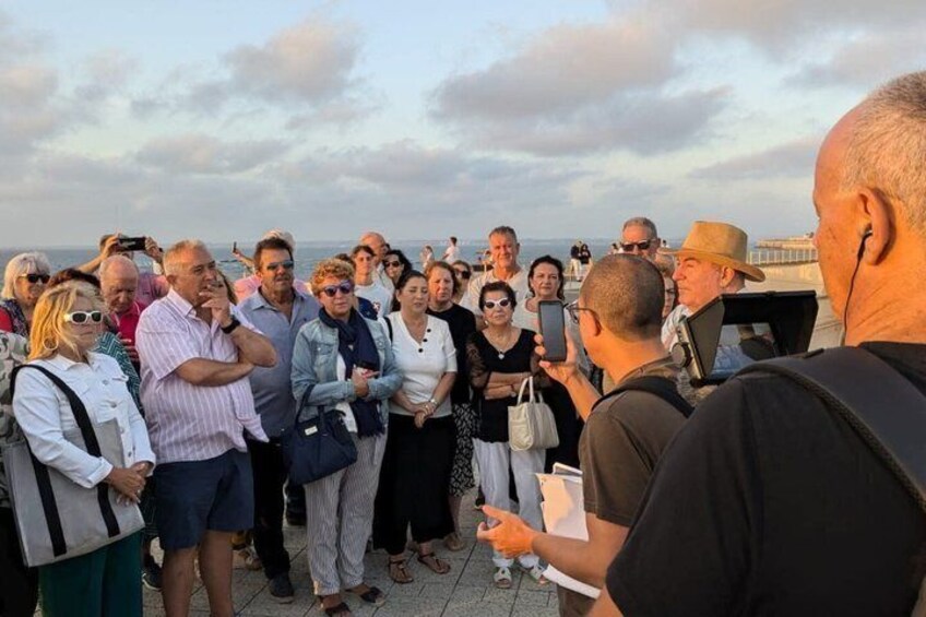 A group of people standing on a pier, some holding cameras, with a body of water and a cloudy sky in the background. 3