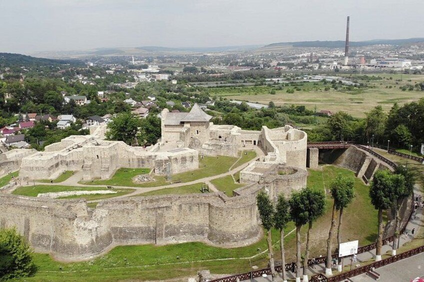 A historic fortification with walls and towers, surrounded by greenery and a distant cityscape. 1