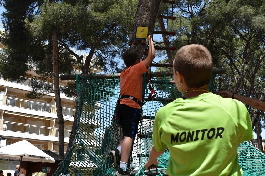 A person climbing a rope ladder in a netted obstacle course with a 'MONITOR' shirt. 1