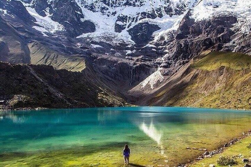 A person standing by a clear turquoise lake surrounded by mountains with snow-capped peaks. 4