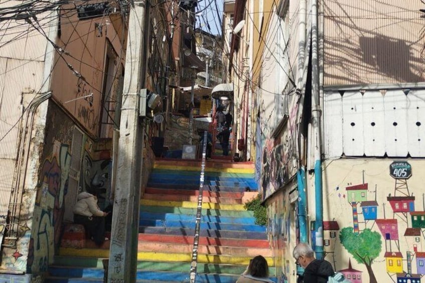 A narrow alley with colorful steps, graffiti, and a mural of houses. 5
