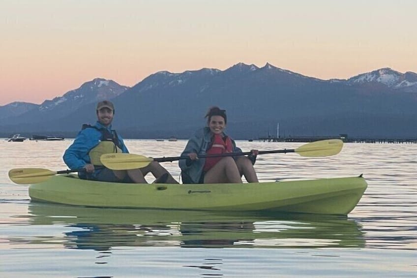 Two people kayaking on calm water with mountains in the background. 1