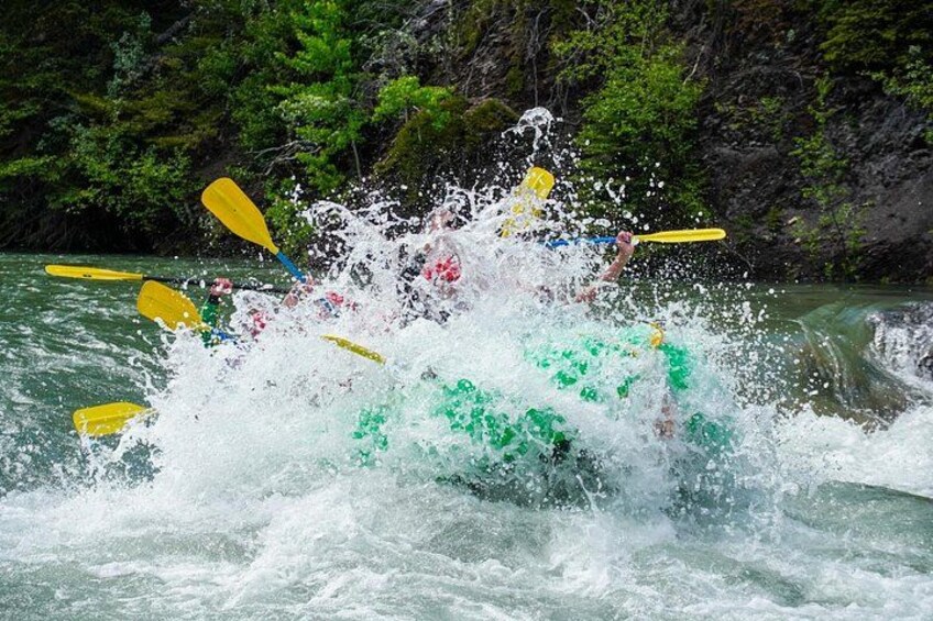 A white water rafting activity with a group of people in a green raft, surrounded by turbulent water and dense green foliage in the background. 3
