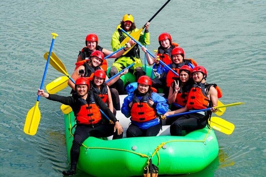 A group of people in a green inflatable raft on water, wearing life jackets and helmets, paddling with yellow paddles. 2