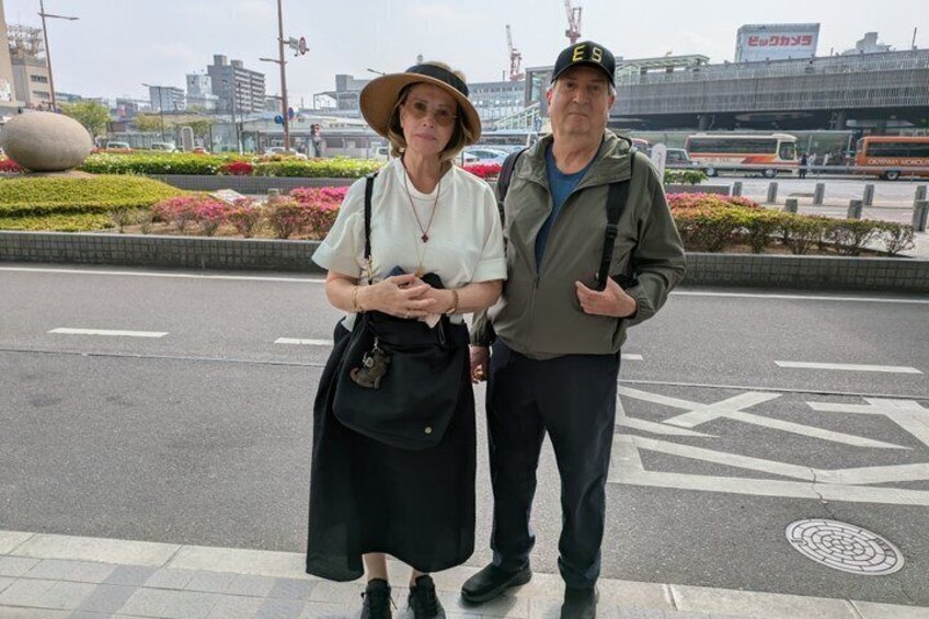 Two people standing on a city street with a roundabout and buildings in the background. 3