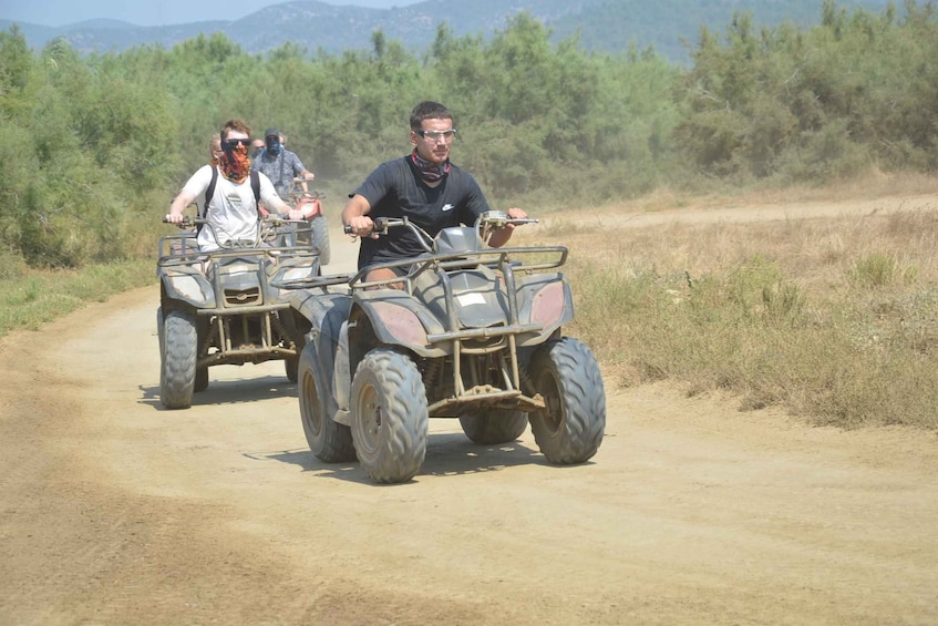 Two people riding atv's on a dirt road with sparse vegetation and mountains in the background. 3