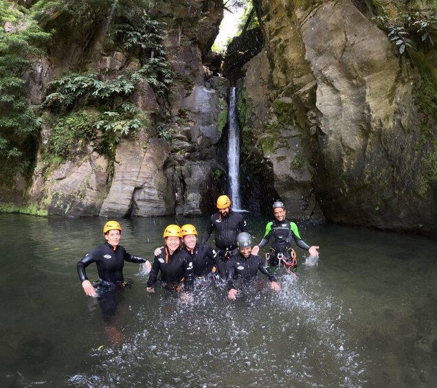 A group of people in wetsuits and helmets standing in a river with a waterfall in the background. 1
