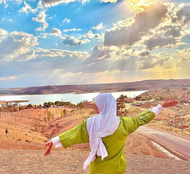 A person with arms outstretched, overlooking a landscape with a body of water and distant hills. 3