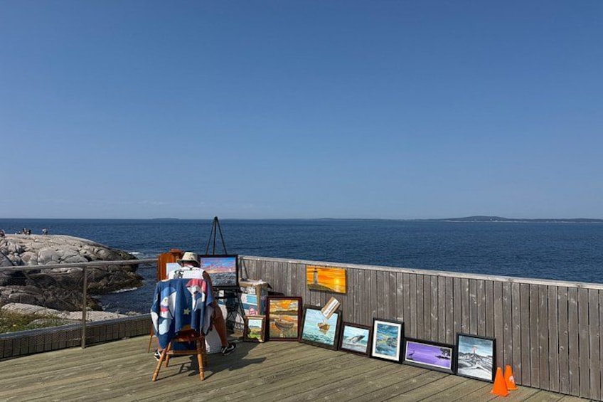A person painting on a wooden deck by the sea, with several framed artworks displayed. 5