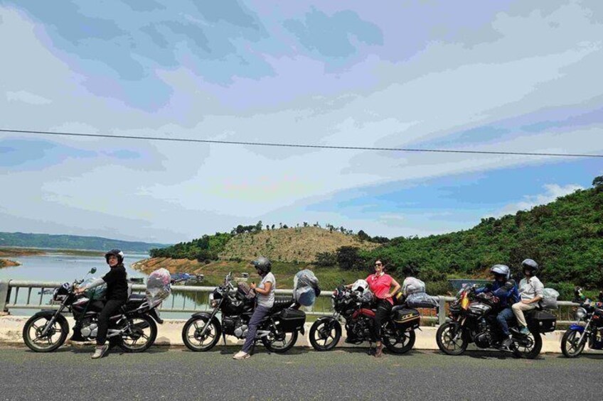A group of motorcyclists parked along a roadside with a hilly landscape and a body of water in the background. 1
