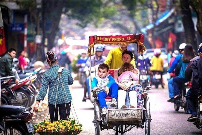 A street scene with a rickshaw carrying passengers, a person pushing the rickshaw, and a basket of fruits. 4