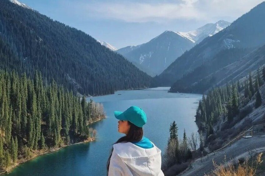 A person in a white jacket and turquoise cap overlooking a lake surrounded by dense forests and snow-capped mountains. 3