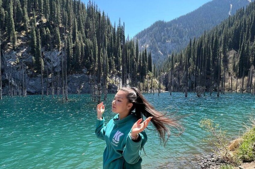 A person standing by a clear turquoise lake surrounded by dense forest. 1