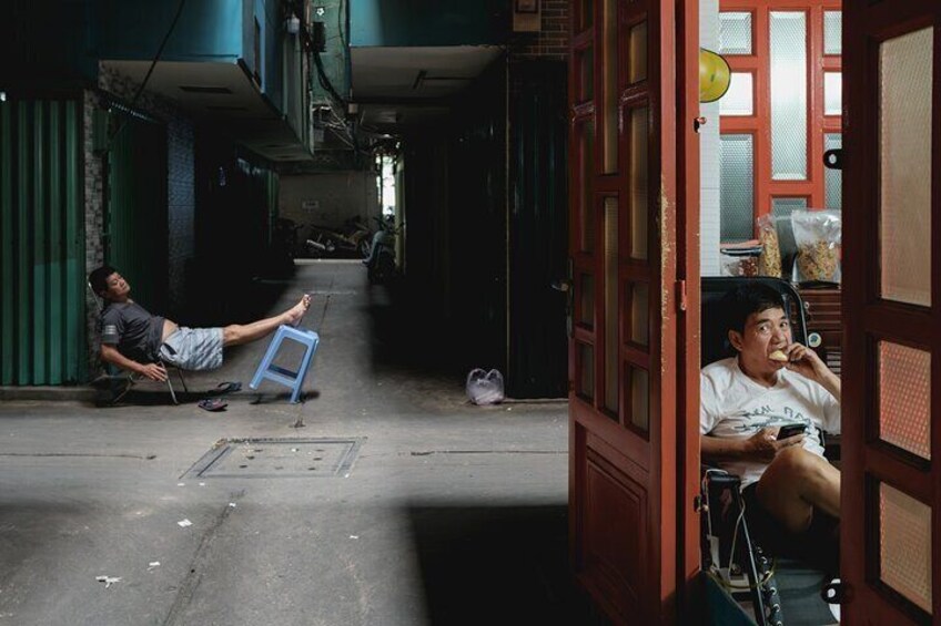 A narrow alleyway with two individuals sitting on the ground, one on a blue stool and the other on a chair. 1