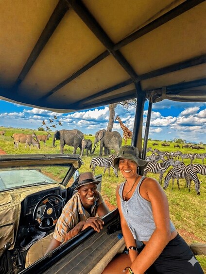A safari vehicle with a driver and a passenger, surrounded by wildlife including zebras, elephants, and giraffes. 2