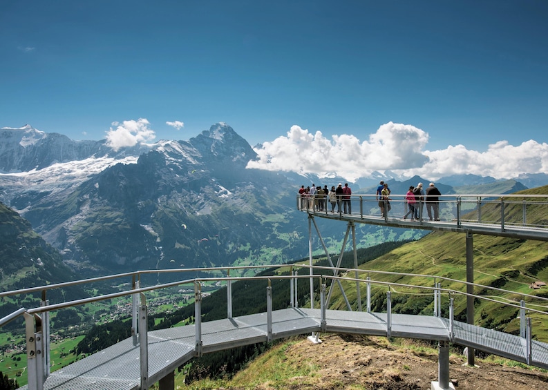 A group of people on a metal walkway overlooking a mountainous landscape with snow-capped peaks and a clear blue sky. 1