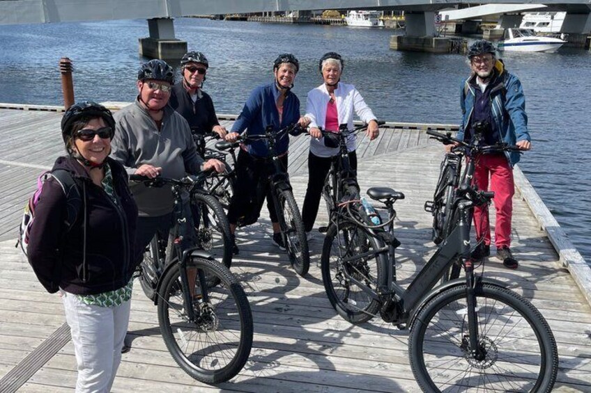 A group of people on a wooden dock by a body of water, with bicycles and a boat in the background. 3