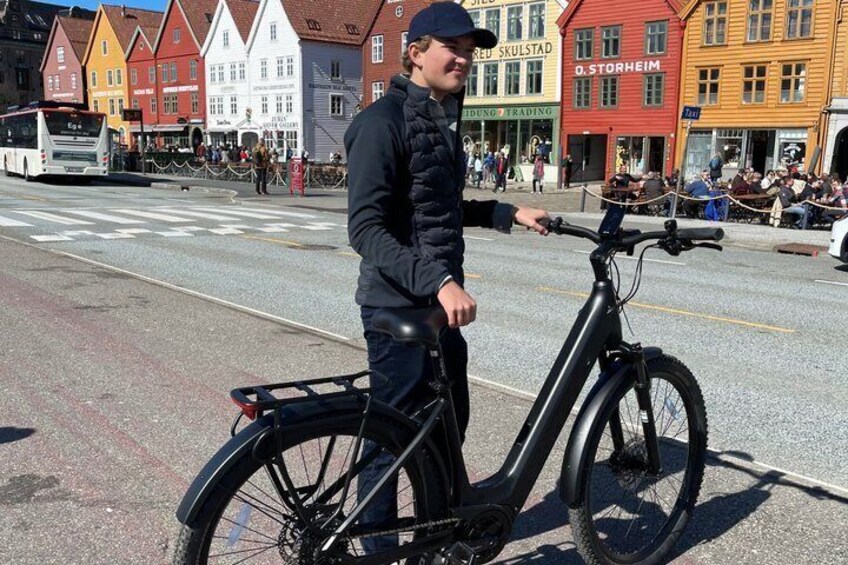 A person standing next to a stationary electric bike on a street with colorful buildings in the background. 5
