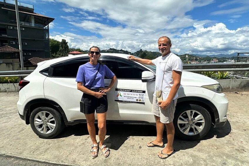 Two people standing next to a white car with a sign on the back. 3