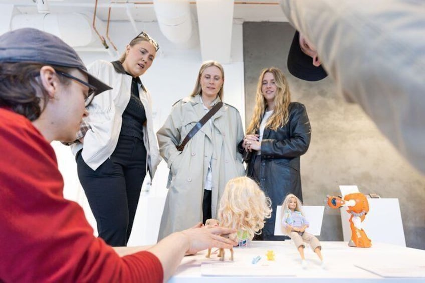 A group of people at an indoor event, some standing and some seated, with a table displaying various items including figurines and a small orange toy. 5