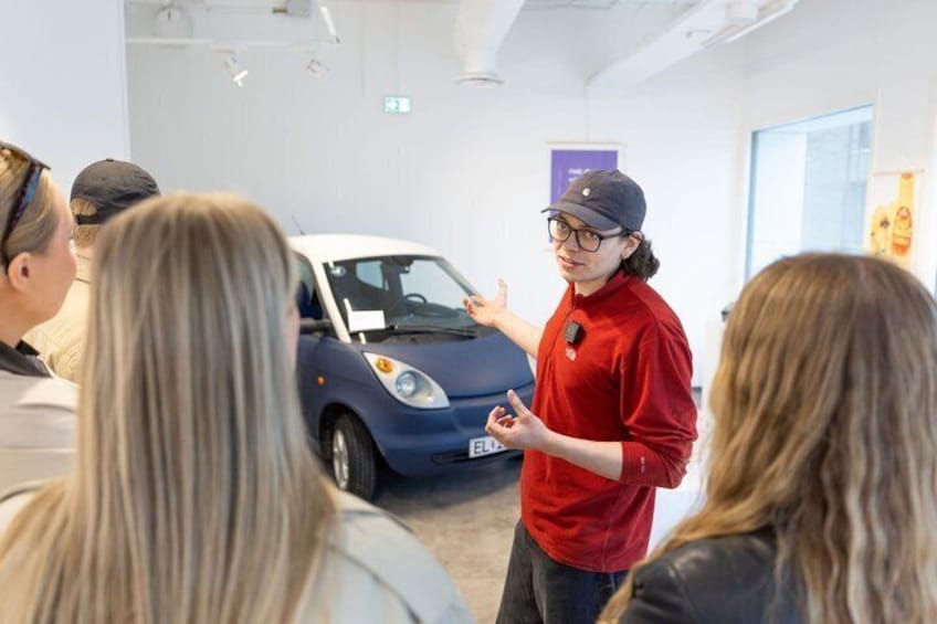 A person in a red shirt and cap standing next to a blue car in an indoor setting with other people. 4