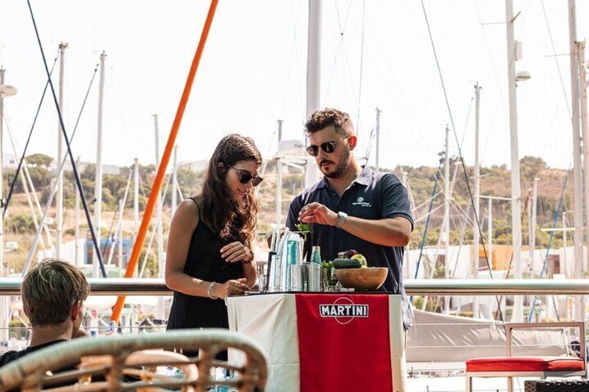 A person preparing drinks at a table with a 'MARTINI' banner, a boat, and a marina in the background. 5