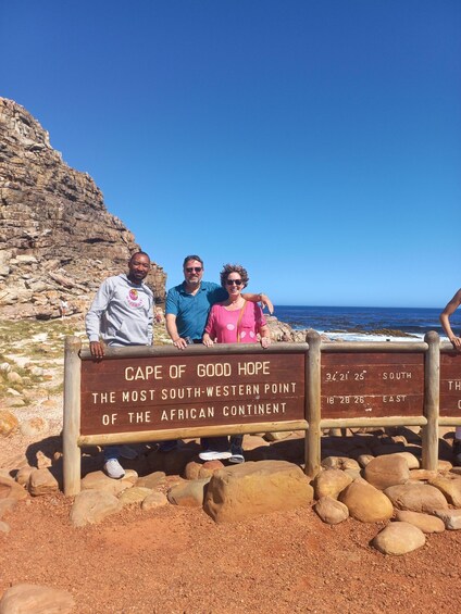 Three people standing in front of a sign at Cape of Good Hope, with a rocky landscape and ocean in the background. 4