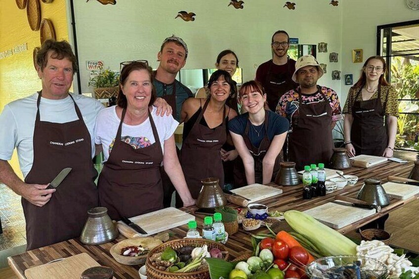 A group of people in aprons standing around a kitchen counter with cooking utensils and fresh produce. 1