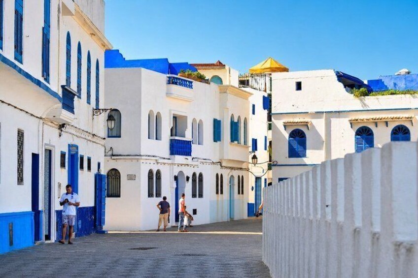 A narrow street with white buildings and blue accents, a clear sky, and a few people walking. 3