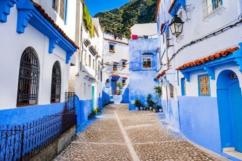 A narrow cobblestone street flanked by buildings painted in white and blue, with arched windows and a mountainous backdrop. 1