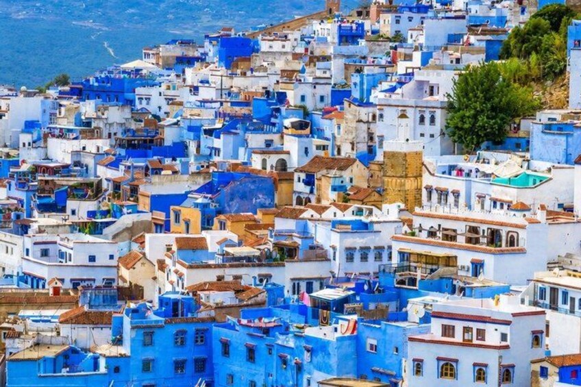A densely packed town with buildings predominantly painted in blue and white, with a hilly backdrop and a body of water in the distance. 2