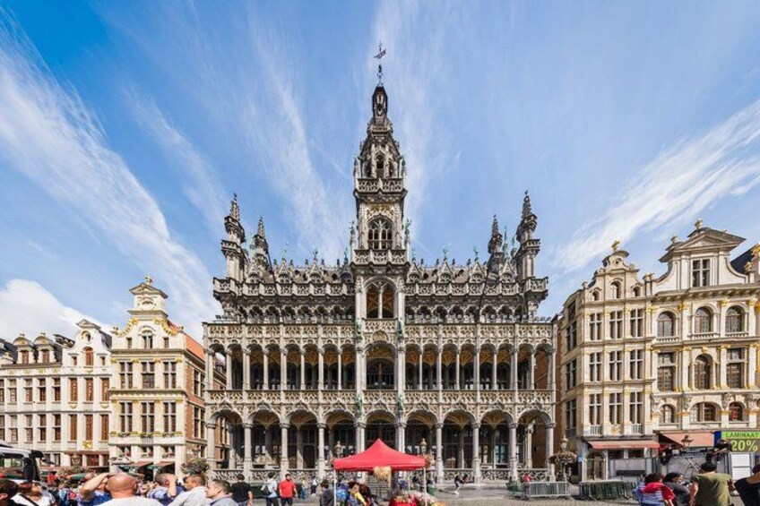A grand, ornate building with intricate architectural details, a prominent central spire, and a surrounding plaza with people and a red canopy. 3