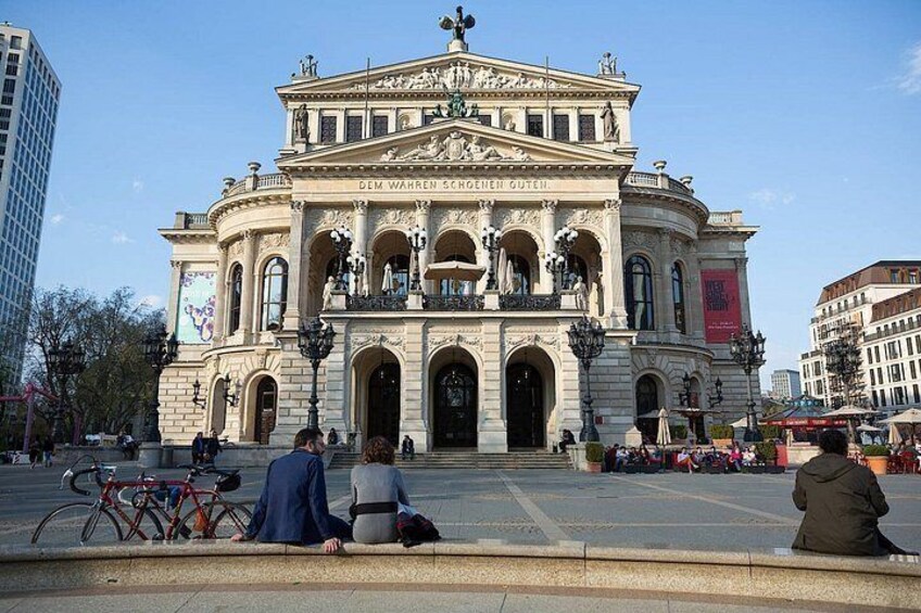 A historic building with ornate architecture, a large open plaza in front, and people sitting and walking around. 4