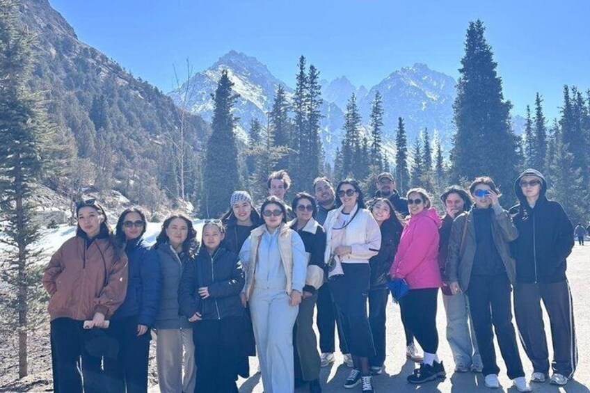 A group of people standing in a snowy mountainous area with trees and mountains in the background. 2