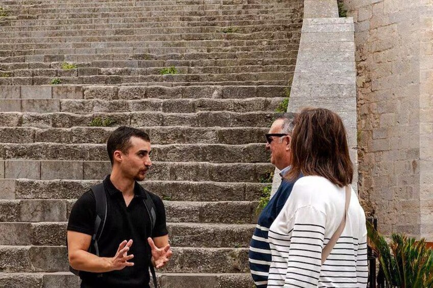 A man gesturing, a woman standing, and a man in a suit, all in front of stone steps. 5