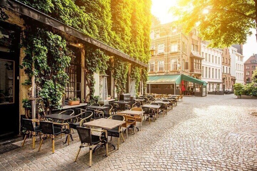 A cobblestone street lined with outdoor cafe tables and chairs, covered by a green awning, with buildings in the background. 2