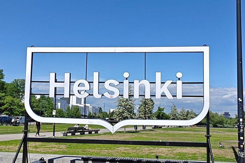 A sign with the word 'Helsinki' in a stylized font, set against a clear blue sky with a cityscape in the background. 2