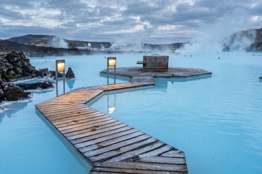 A geothermal spa with wooden walkways and benches, surrounded by rocky terrain and steam rising from the water. 2