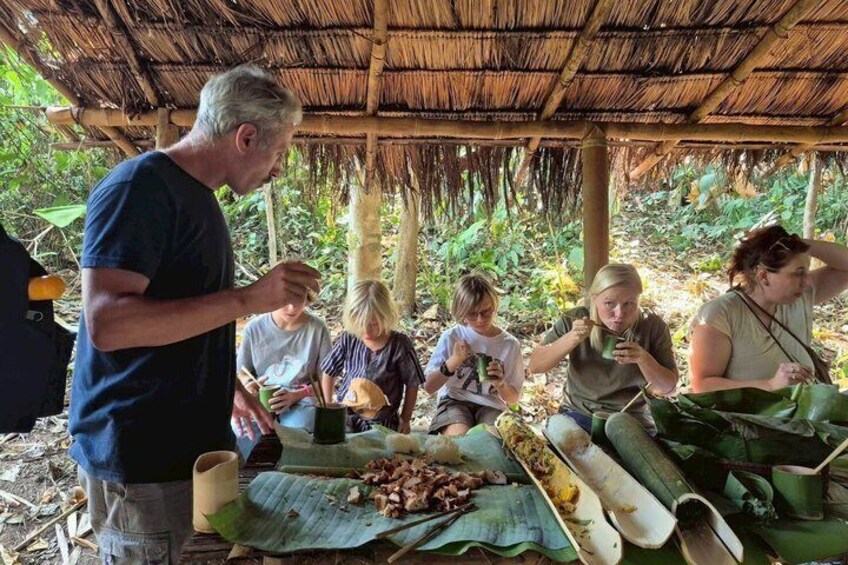 A group of people eating under a thatched roof, surrounded by greenery. 3