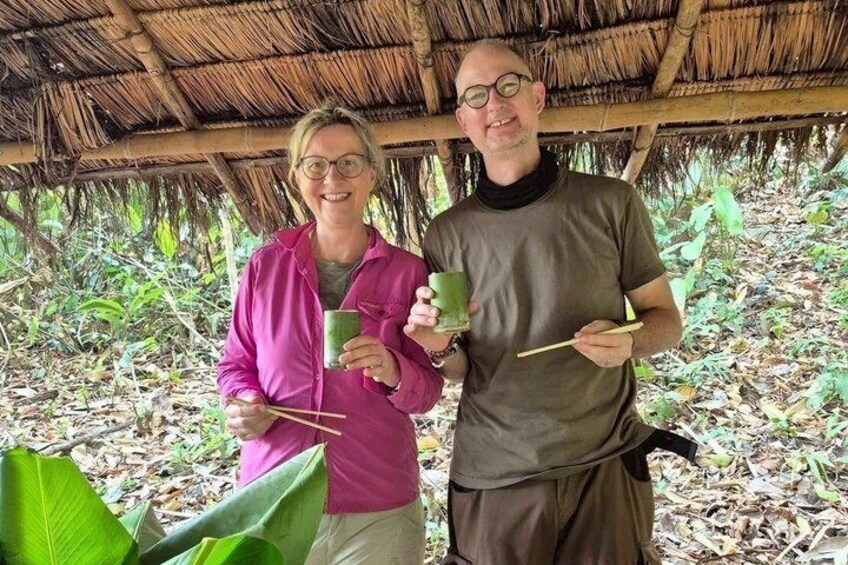 Two people holding green drinks and chopsticks, standing under a thatched roof structure surrounded by foliage. 2