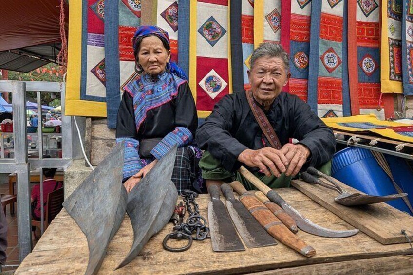 Two individuals seated at a table with traditional tools and colorful textiles in the background. 2