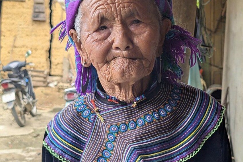 An elderly person wearing a colorful traditional garment with intricate patterns, standing in front of a rustic building with a motorcycle parked nearby. 5