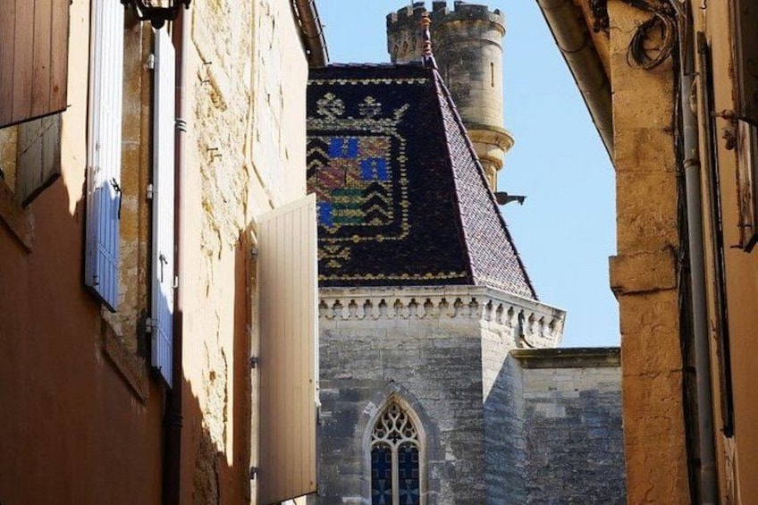 A narrow street with a medieval building featuring a tower and a patterned roof. 2