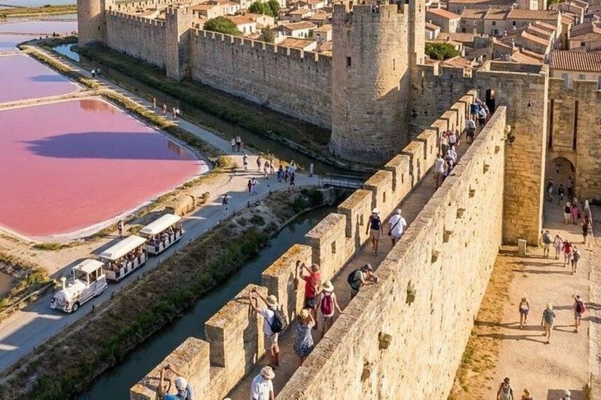 A historic stone wall with people walking along it, a body of water with pinkish hue, and a castle in the background. 3