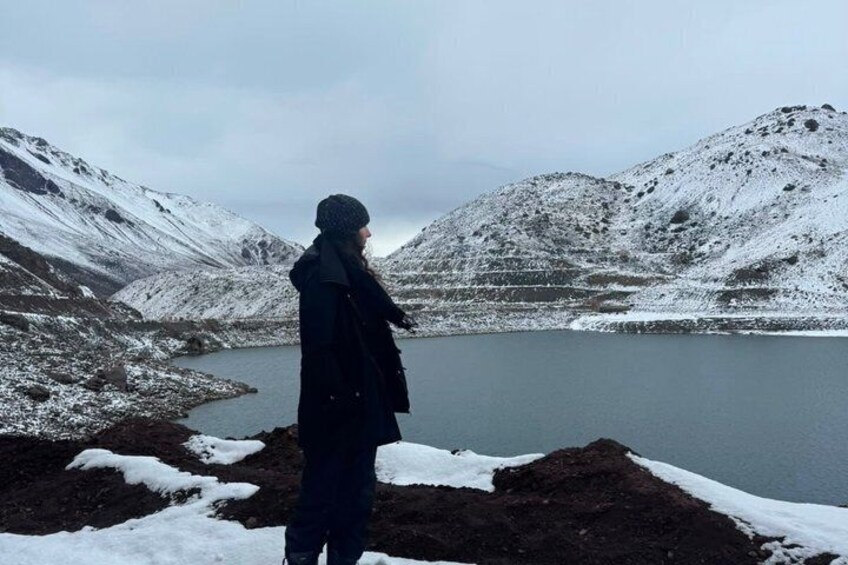 A person standing by a snow-covered lake with mountains in the background. 1