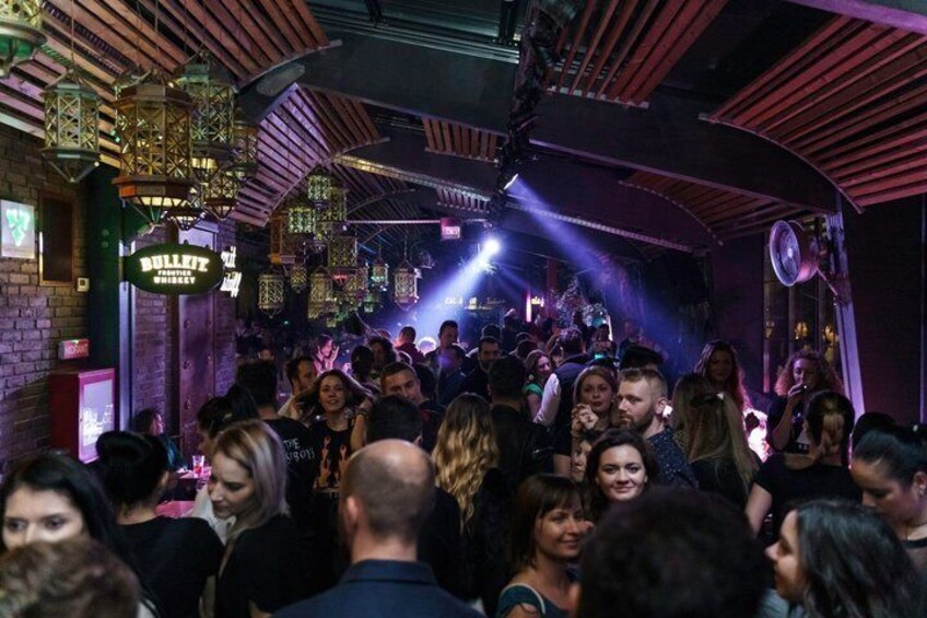 A crowded bar with a stage, hanging lanterns, and a sign reading 'Bullitt Whiskey.' 5